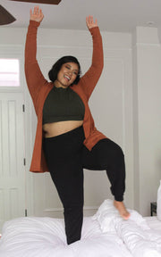 Plus size model jumping on bed in joggers, cropped tank, and cardigan